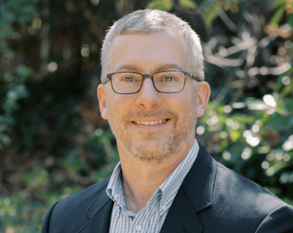 A man with short gray hair, glasses, and a beard smiles at the camera. He is wearing a dark blazer over a striped shirt. The background features greenery and dappled sunlight.